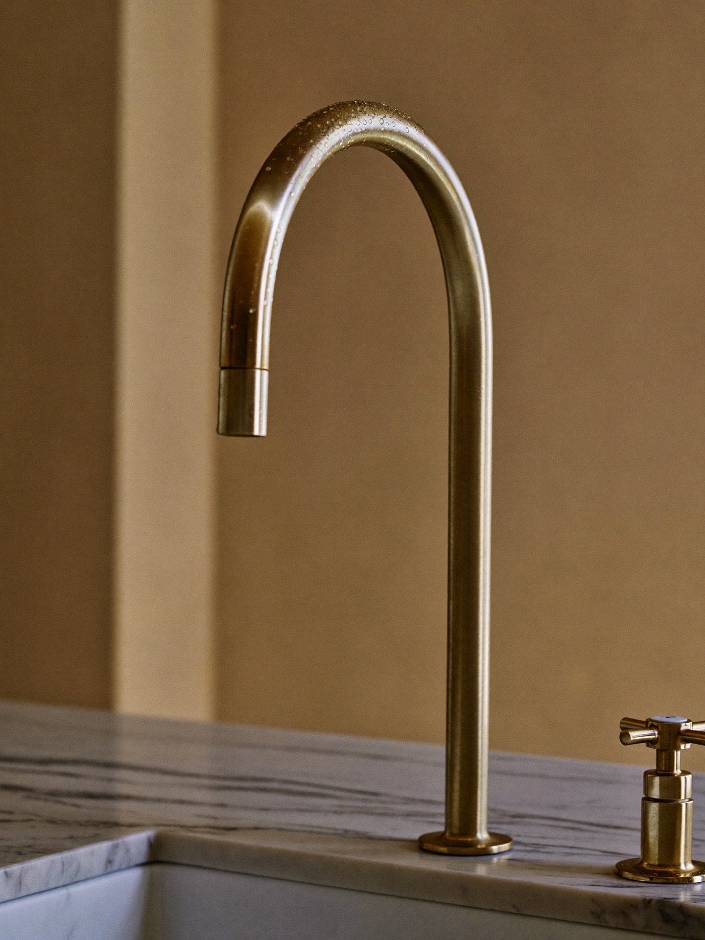 Unlacquered brass gooseneck faucet beaded with water over a honed Calacatta marble counter, warm plaster wall behind