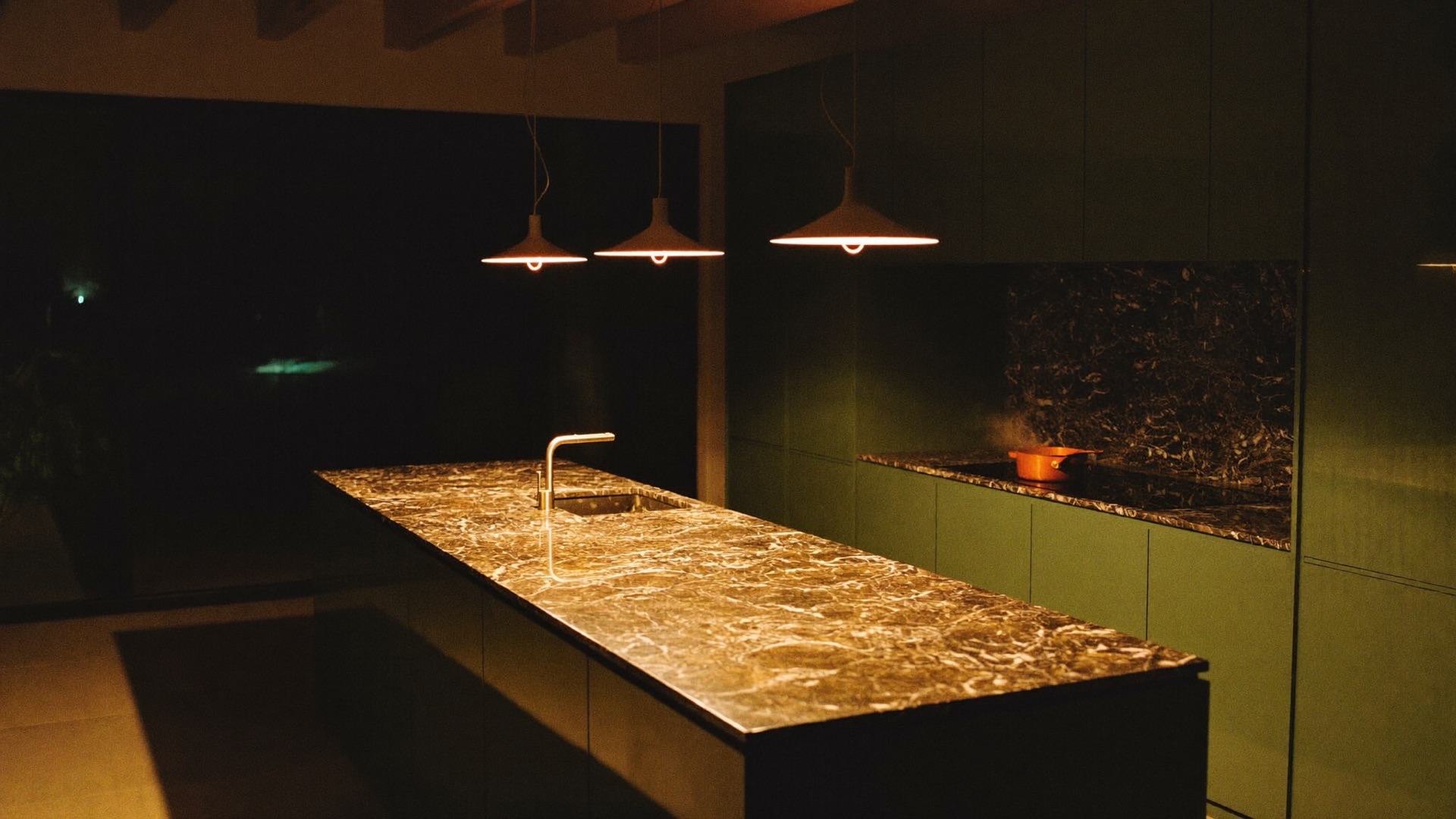 A dark green kitchen after dusk, three pendant lamps pooling warm light on a marble island, a copper pan steaming on the range